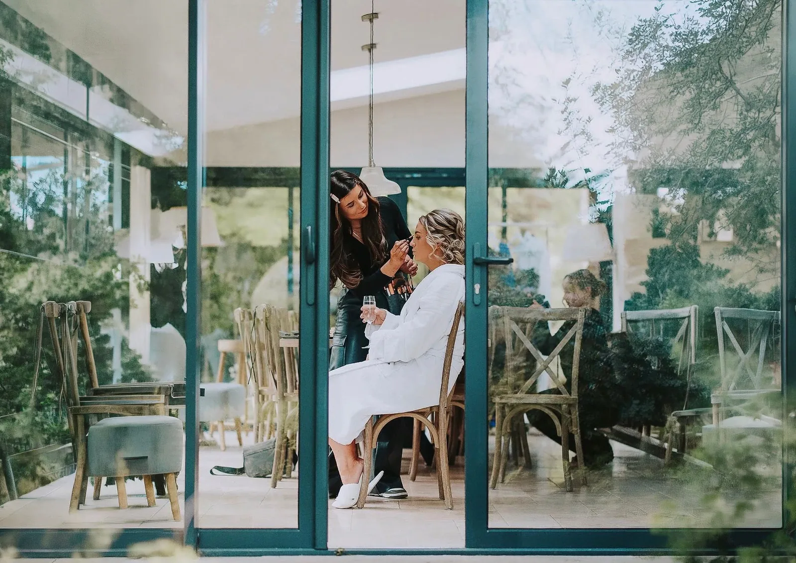 Chelsea applying makeup to a bride in a robe, seen through glass doors